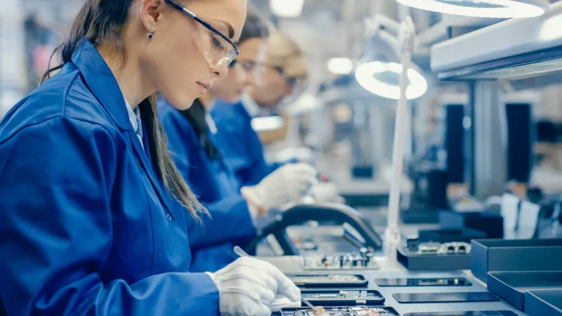 female electronics factory workers in blue work coat and protective glasses assembling printed circuit boards for smartphones with tweezers high tech factory with more employees in the background