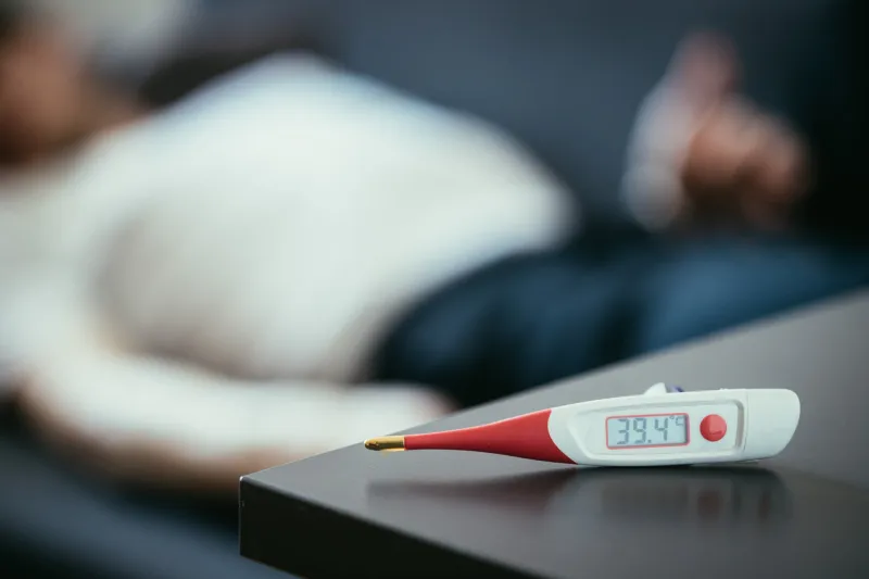 fever thermometer in the foreground, ill man lying on the sofa in the blurry background