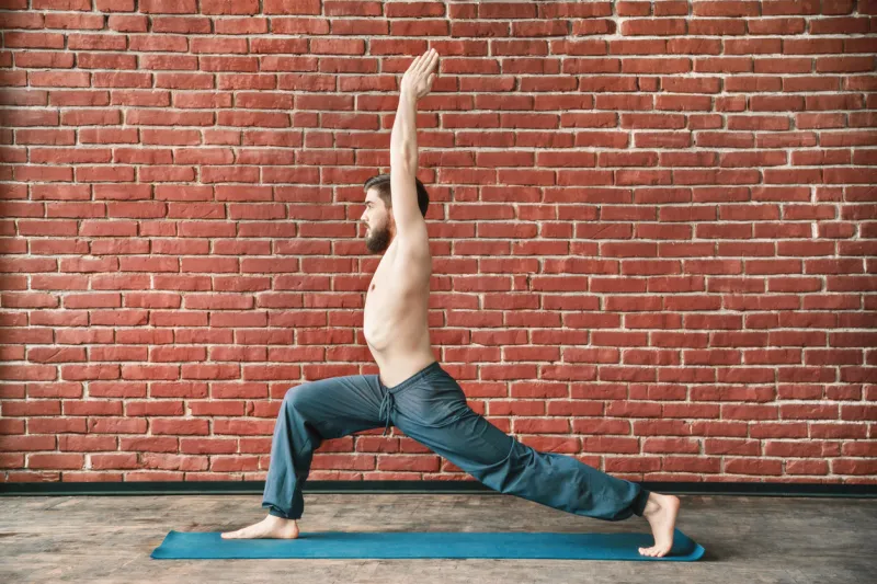 attractive man with dark hair and beard wearing trousers doing yoga warrior position on blue matt at wall background, copy space, portrait, virabhadrasana asana