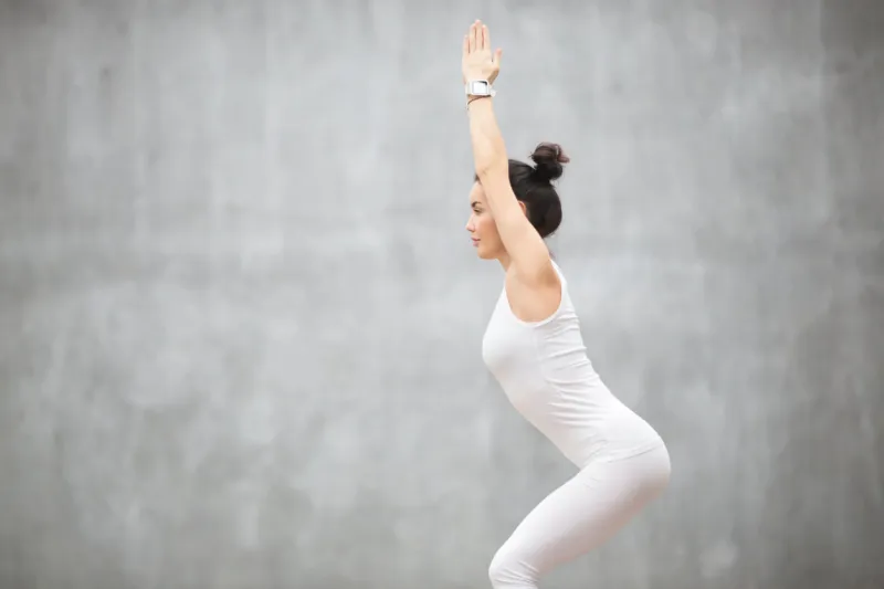 side view portrait of beautiful young woman wearing white sportswear working out against grey wall, doing yoga or pilates exercise standing in chair, utkatasana pose copy space