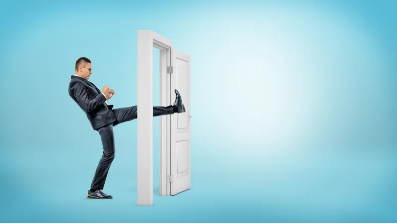 a businessman in side view kicks a small white door open with his leg on blue backgrounds business and success opening all doors aggressive business approach