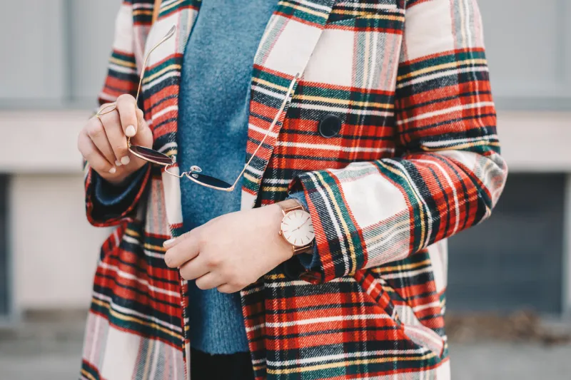 close-up detail of fashion accessories, stylish young woman wearing an overcoat with a tartan pattern, a golden ring, a pair of modern sunglasses and a wrist watch