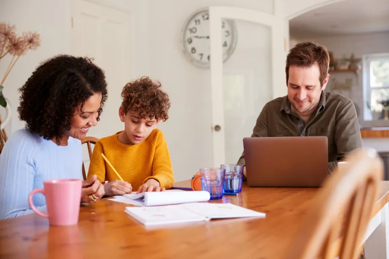 father works on laptop as mother helps son with homework on kitchen table