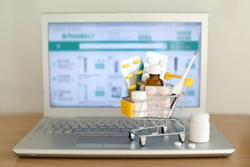shopping cart toy with medicaments in front of laptop screen with pharmacy web site on it pills, blister packs, medical bottles, thermometer set health care and internet shopping