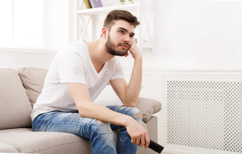 young man watching television, using remote control to switch channels guy bored with what he sees on tv screen sitting on couch in living room at home, copy space
