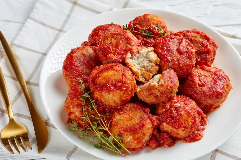 fried fish balls in tomato sauce on a white plate on a wooden table with golden fork and knife