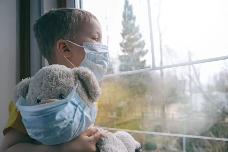 sad illness child on home quarantine boy and his teddy bear both in protective medical masks sits on windowsill and looks out window virus protection, coronavirus pandemic, prevention epidemic