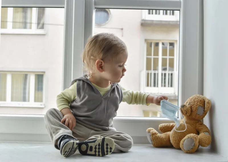 child in home quarantine at the window putting a medical mask on his sick teddy bear, for protection against viruses during coronavirus covid-19 and flu outbreak children and illness disease concept