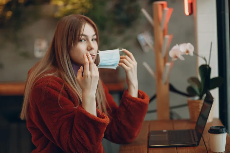 young woman sits at a table in cafe and putting on medical mask