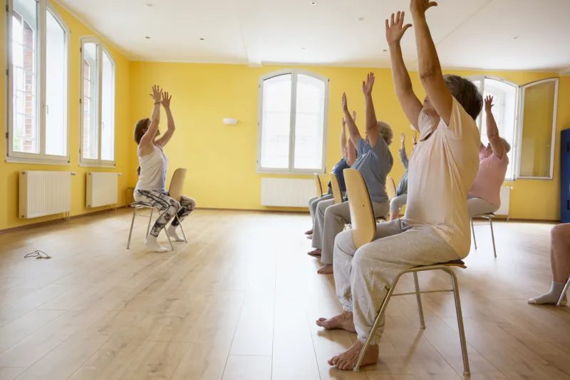 teacher and active senior women yoga class on chairs, arms raised