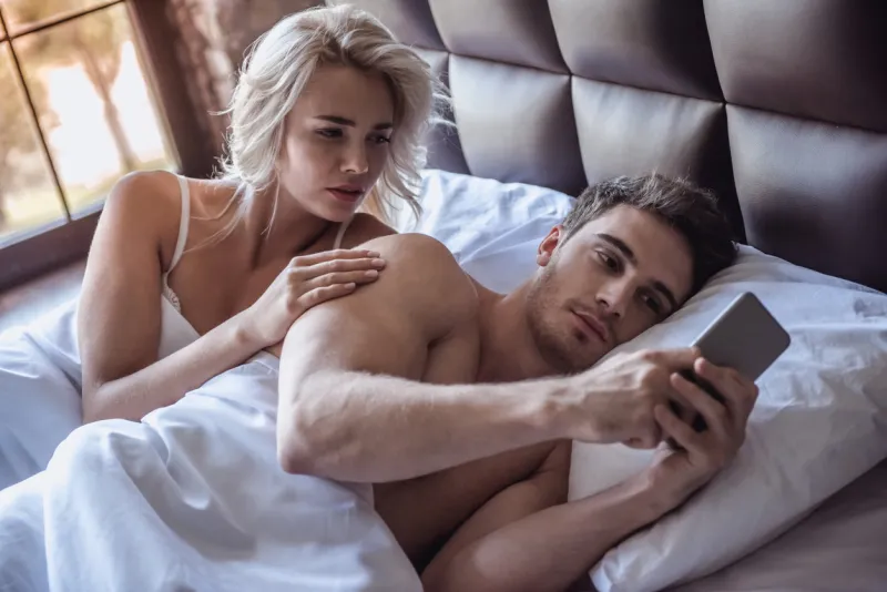 beautiful young couple is lying in bed, guy is using a smart phone, girl is looking with reproach