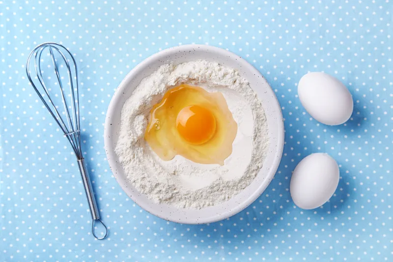 broken egg yolk and flour in a bowl, whisk and white eggs on the side food preparation concept baking ingredients top view