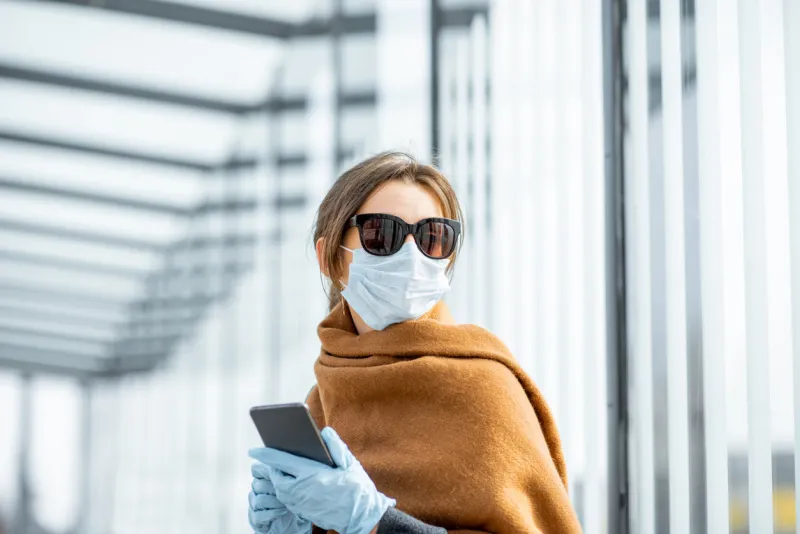 young woman in face mask, medical gloves and sunglasses with a smart phone at the public transport stop during an epidemic concept of social distance and online communication