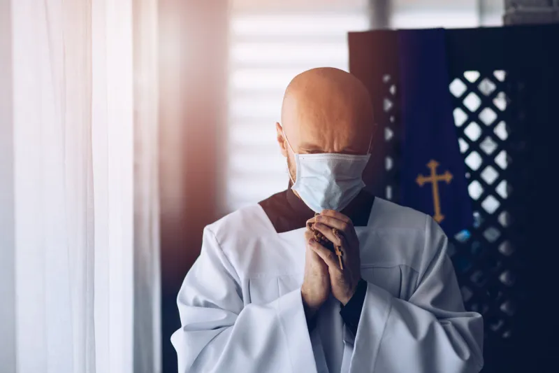man priest in medical mask praying with cross and rosary church during corona virus epidemic