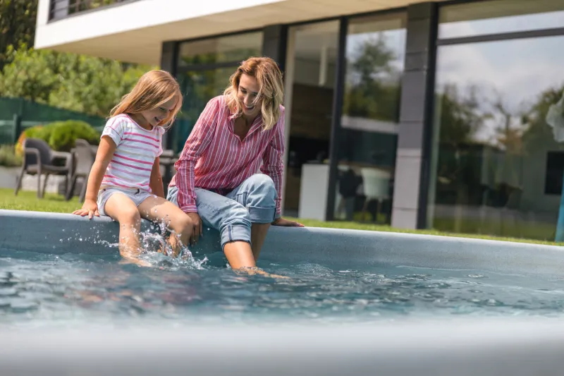 mirthful woman and her daughter putting legs into the water of swimming pool outdoors