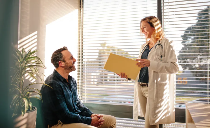 smiling female doctor holding a medical report file and talking with male patient sitting on sofa at hospital reception doctor sharing good medical test results with the patient
