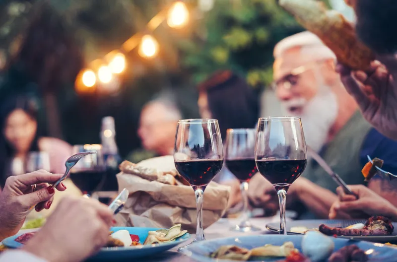 happy family eating and drinking red wine at dinner barbecue party outdoor - mature and young people dining together on rooftop - youth and elderly weekend lifestyle activities - focus on wineglass