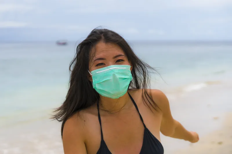 use of medical face mask in public places- young attractive asian korean woman enjoying beach holidays wearing bikini and protective facial mask in prevention vs virus infection