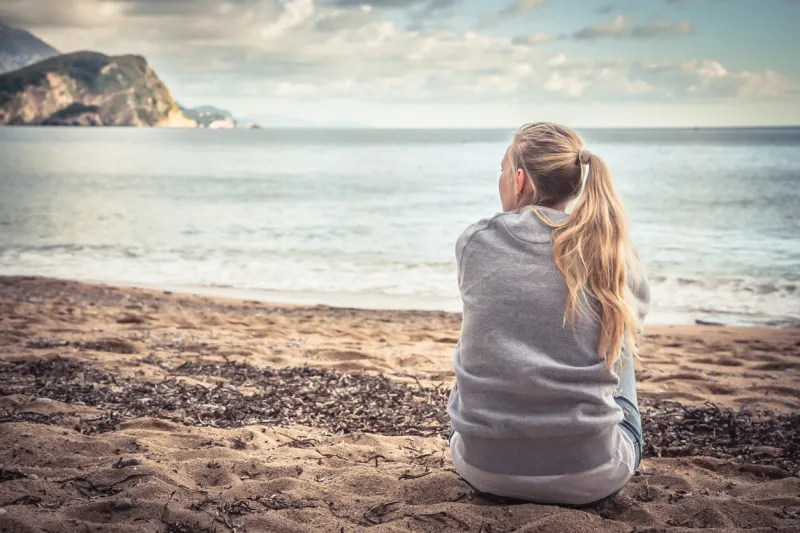pensive lonely young woman tourist sitting on beach hugging her knees and looking into the distance with hope