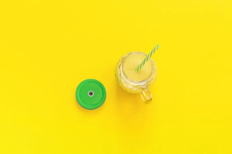 glass jar with natural yellow fruit juice, smoothie or fresh with green lid and cocktail straw on yellow background top view copy space template