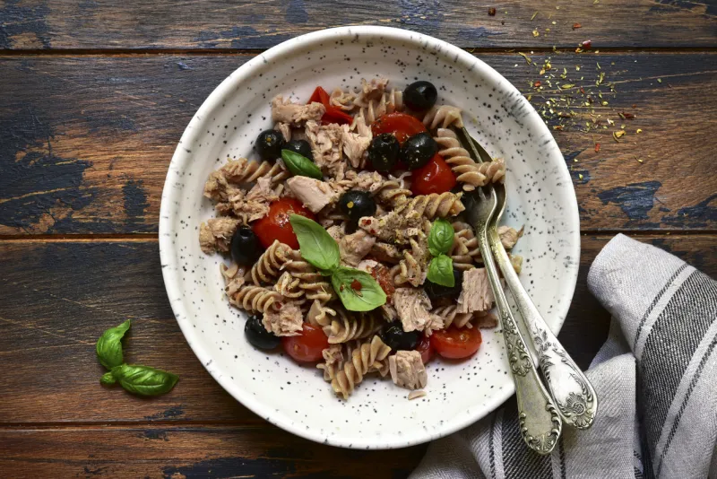 whole wheat pasta fusilli with tuna, tomato and black olives in a white bowl on a dark wooden background top view with copy space