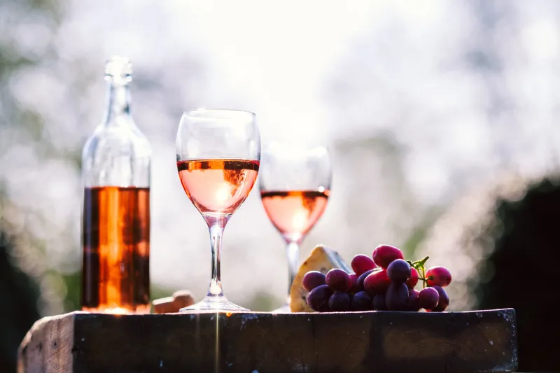 rose wine filled bottle,glasses and a cork on a large cheese board with red grapes and stilton cheese in a natural sun light environment with a garden background with copy space