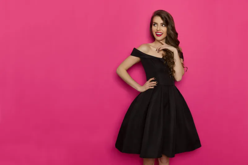 beautiful young woman in elegant black cocktail dress is holding hand on chin, looking away and thinking three quarter length studio shot on pink background
