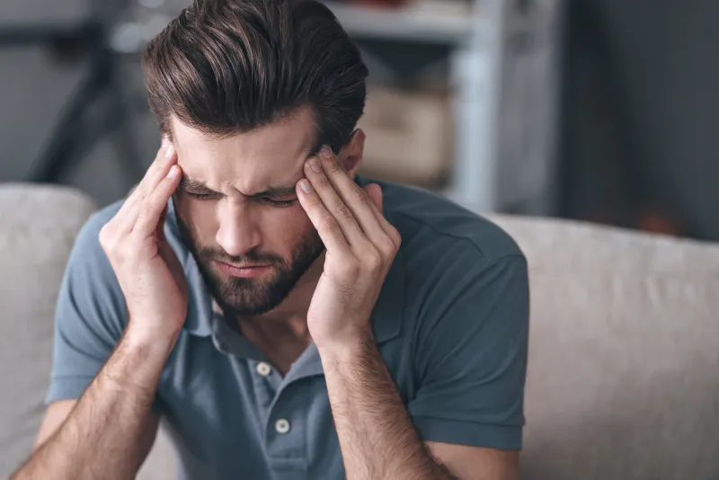 frustrated handsome young man touching his head and keeping eyes closed while sitting on the couch at home