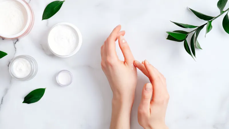 cosmetic cream on female hands, jars with milk swirl cream and green leaves on white marble table flat lay, top view woman applying organic moisturizing hand cream hand skin care concept