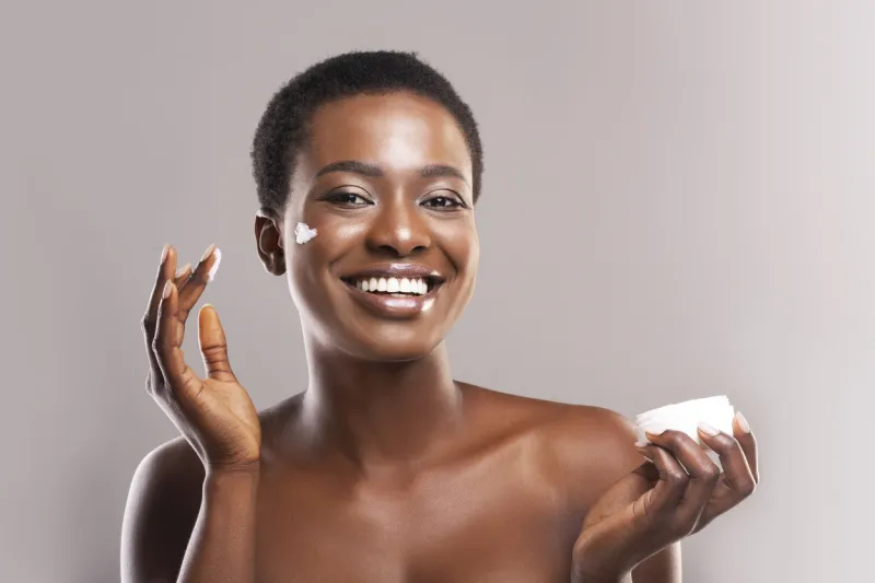 skin hydration portrait of cheerful black girl holding and applying moisturizing cream on face, grey background with free space
