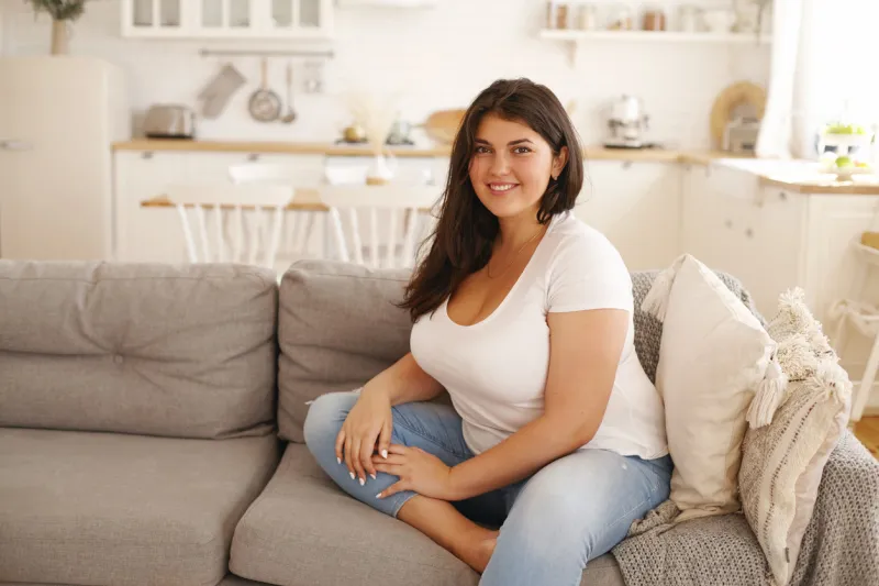 relaxation, leisure, rest, domesticity and coziness beautiful charming young female with chubby cheeks and curvy body sitting on sofa barefooted, keeping one foot on floor, smiling at camera