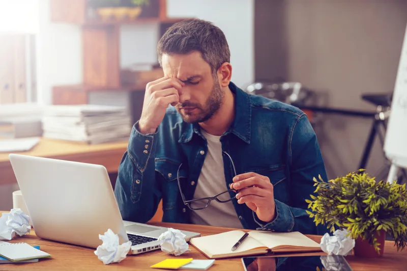 frustrated young man massaging his nose and keeping eyes closed while sitting at his working place in office