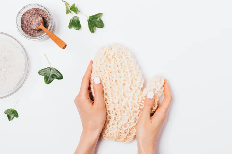 woman's hands holding natural massage mitten washcloth next to coffee body scrub and bowl of sea salt on white background, top view