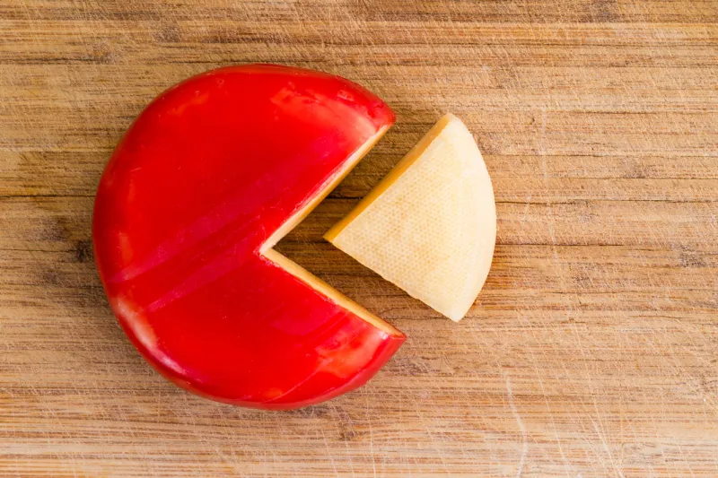 wheel of fresh gouda cheese with a red rind and a single wedge portion cut out, peeled and separated to the side on a wooden cutting board