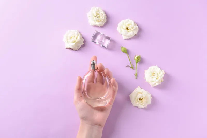 female hand with bottle of perfume and flowers on color background