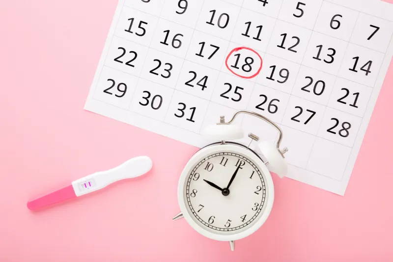 calendar, test with two stripes and white alarm clock on pastel pink table background positive result pregnancy time beginning closeup top down view