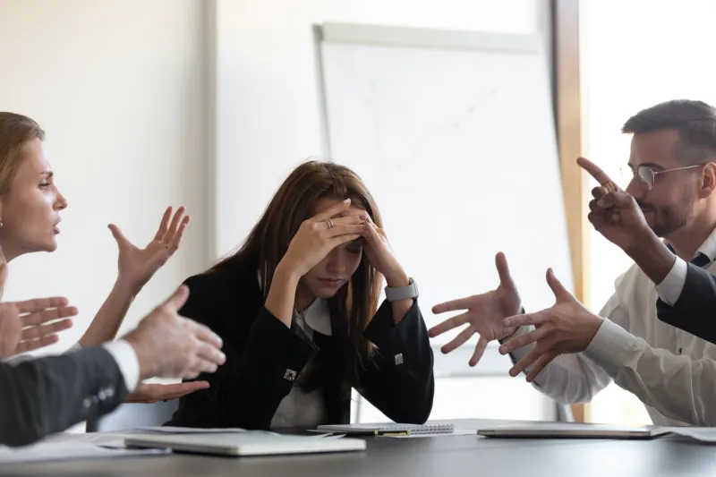 frustrated millennial female worker sitting at table with colleagues, felling tired of working quarreling at business meeting upset stressed young businesswoman suffering from head ache at office