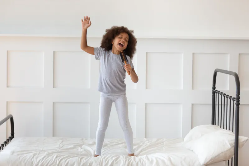 cute little preschooler african american girl stand on bed sing in toy hairbrush microphone, funny small artistic biracial kid have fun playing at home in morning, perform entertain in bedroom