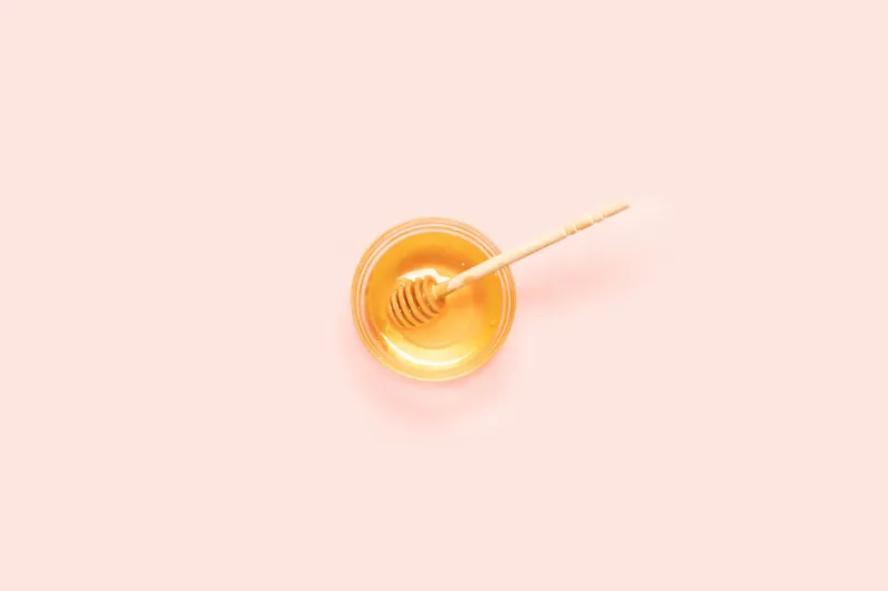 top view of fresh honey in glass bowl with wooden spoon on pink background