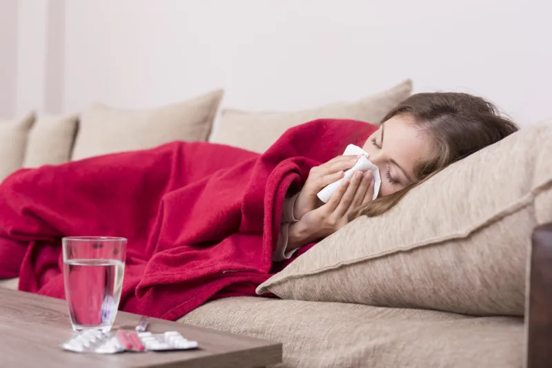 sick woman covered with a blanket lying in bed with high fever and a flu, blowing her nose pills and glass of water on the table