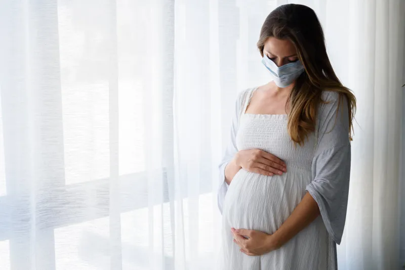 pregnant woman standing by the big window with face medical mask on worries about child birth during pandemic