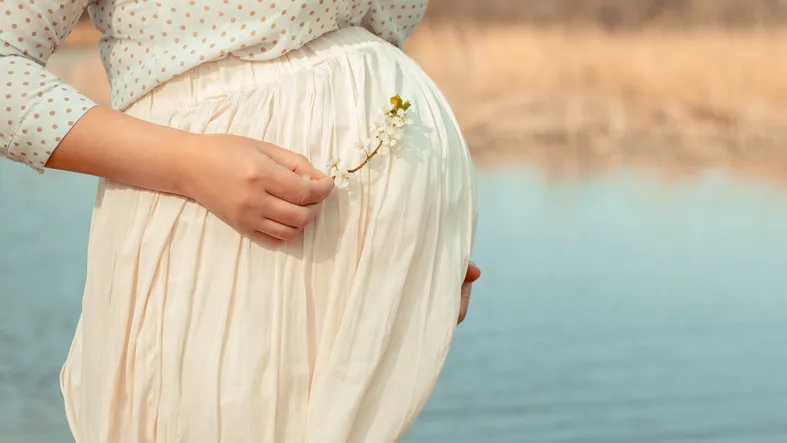 pregnant girl in a white dress on a background of a river he hugs his stomach with his hands and holds a flowering branch of a spring tree vintage toning, focus on pregnant belly pregnancy planning