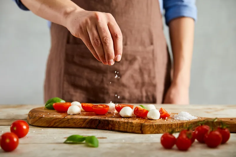male cook is adding a pinch of salt on tomatoes, mozzarella and basil on a olive cutting board