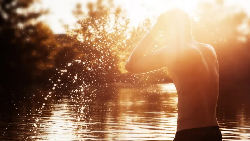 a young man is standing in a river and washing his face against the sunset a man with a bare torso is standing in the water