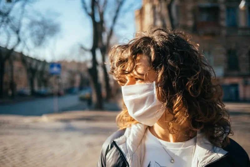young woman in protective face mask looking away while standing on empty street in city on sunny day, copy space