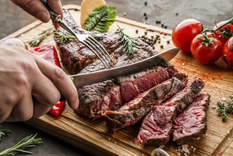 hands cut grilled tomahawk meat medium rare or rib eye steak on wooden cutting board with grilled vegetables on dark background, close up