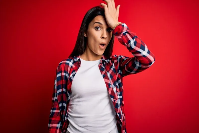 young beautiful brunette woman wearing casual shirt standing over isolated red background surprised with hand on head for mistake, remember error forgot, bad memory concept