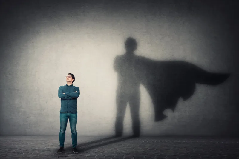 brave man keeps arms crossed, looks confident, casting a superhero with cape shadow on the wall ambition and business success concept leadership hero power, motivation and inner strength symbol