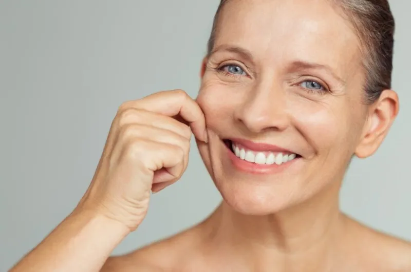 senior woman pulling cheeks to feel softness and looking at camera beauty portrait of happy mature woman smiling with hands on cheek isolated over grey background aging process and perfect skin concept