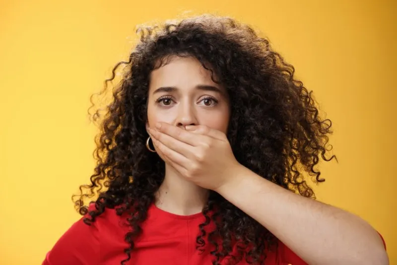 close-up shot of concerned and insecure troubled woman keep silent scare of telling anyone her problem covering mouth with palm not to scream or slip word looking sad and sorrow over yellow background